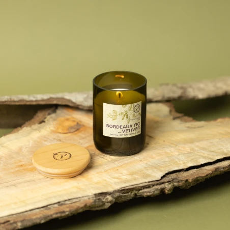 Vela Paddywax Eco Bordeaux Fig & Vetiver encendida sobre corteza de madera, con su tapa de madera grabada al lado