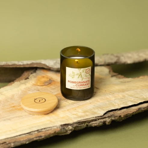 Vela Paddywax Eco Pomegranate & Currant encendida sobre corteza de madera, con su tapa de madera grabada al lado