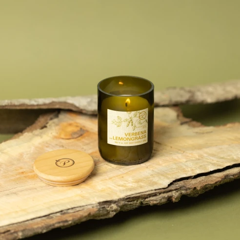 Vela Paddywax Eco Verbena & Lemongrass encendida sobre corteza de madera, con su tapa de madera grabada al lado