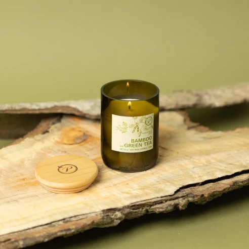 Vela Paddywax Eco Bamboo & Green Tea encendida sobre corteza de madera, con su tapa de madera grabada al lado