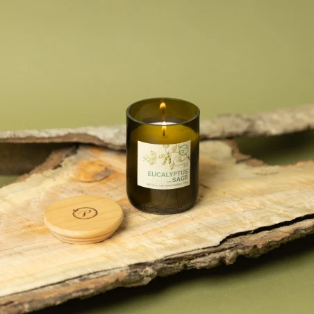 Vela Paddywax Eco Eucalyptus & Sage encendida sobre corteza de madera, con su tapa de madera grabada al lado