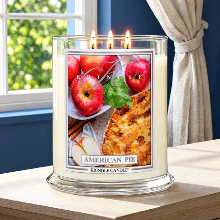 Vela Kringle Candle American Pie de 3 mechas encendida en una mesa de madera junto a una ventana con cortinas de lino