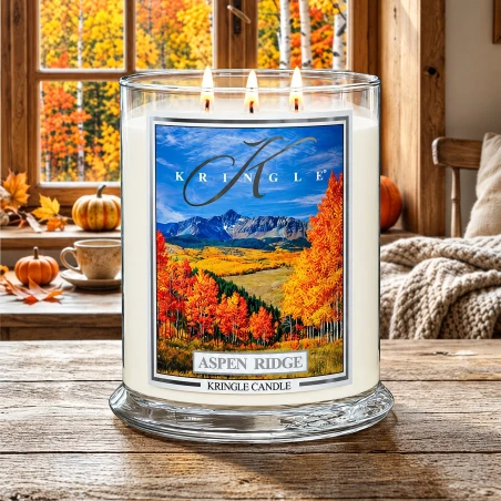 Vela Kringle Candle Aspen Ridge de 3 mechas encendida en una mesa de madera rústica junto a una ventana con bosque otoñal