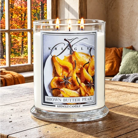 Vela Kringle Candle Brown Butter Pear de 3 mechas encendida en una mesa de madera junto a una ventana con árboles otoñales