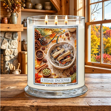 Vela Kringle Candle Cedar Incense de 3 mechas encendida en una cabaña de madera con chimenea de piedra y vistas al otoño
