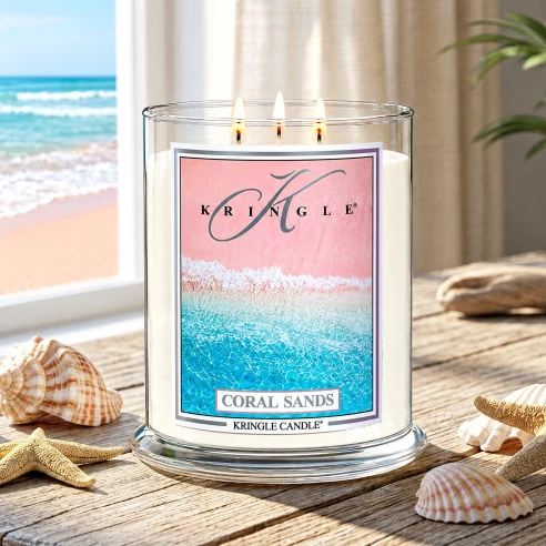 Vela Kringle Candle Coral Sands de 3 mechas encendida en una mesa de madera con conchas junto a una ventana al mar