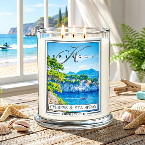 Vela Kringle Candle Cypress & Sea Spray de 3 mechas encendida en una mesa con estrellas de mar y conchas frente a la costa
