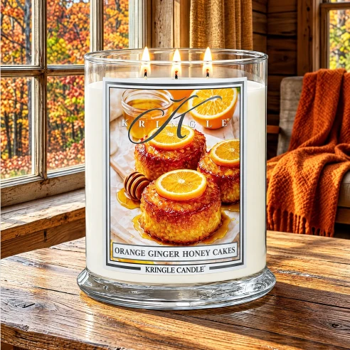 Vela Kringle Candle Orange Ginger Honey Cakes de 3 mechas encendida en una cabaña de madera con ventanales y bosque otoñal