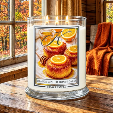 Vela Kringle Candle Orange Ginger Honey Cakes de 3 mechas encendida en una cabaña de madera con ventanales y bosque otoñal